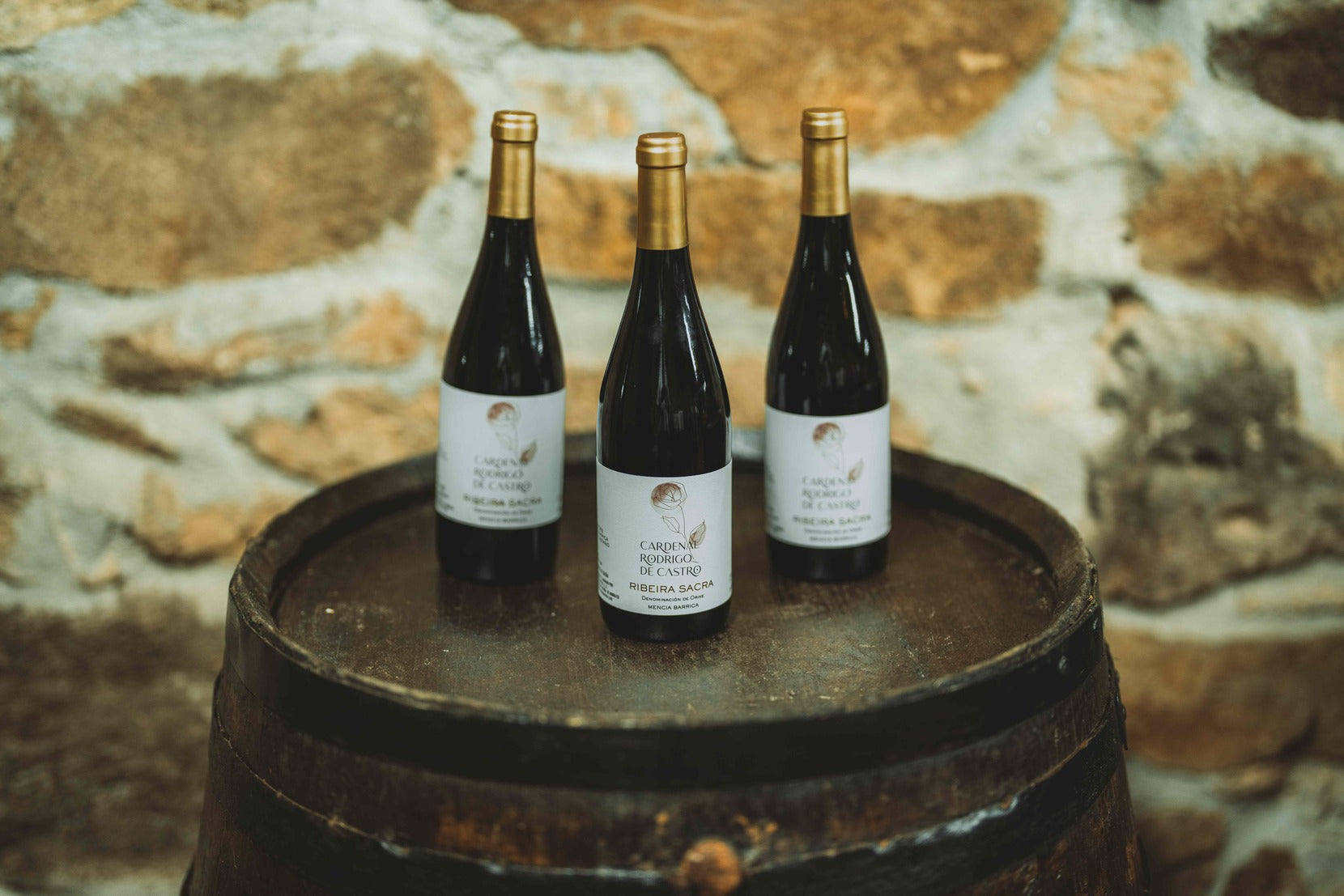 Three bottles of Cardenal Rodrigo de Castro Barrica with gold capsules on an old barrel, against a stone wall