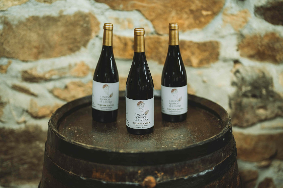 Three bottles of Cardenal Rodrigo de Castro Barrica with gold capsules on an old barrel, against a stone wall